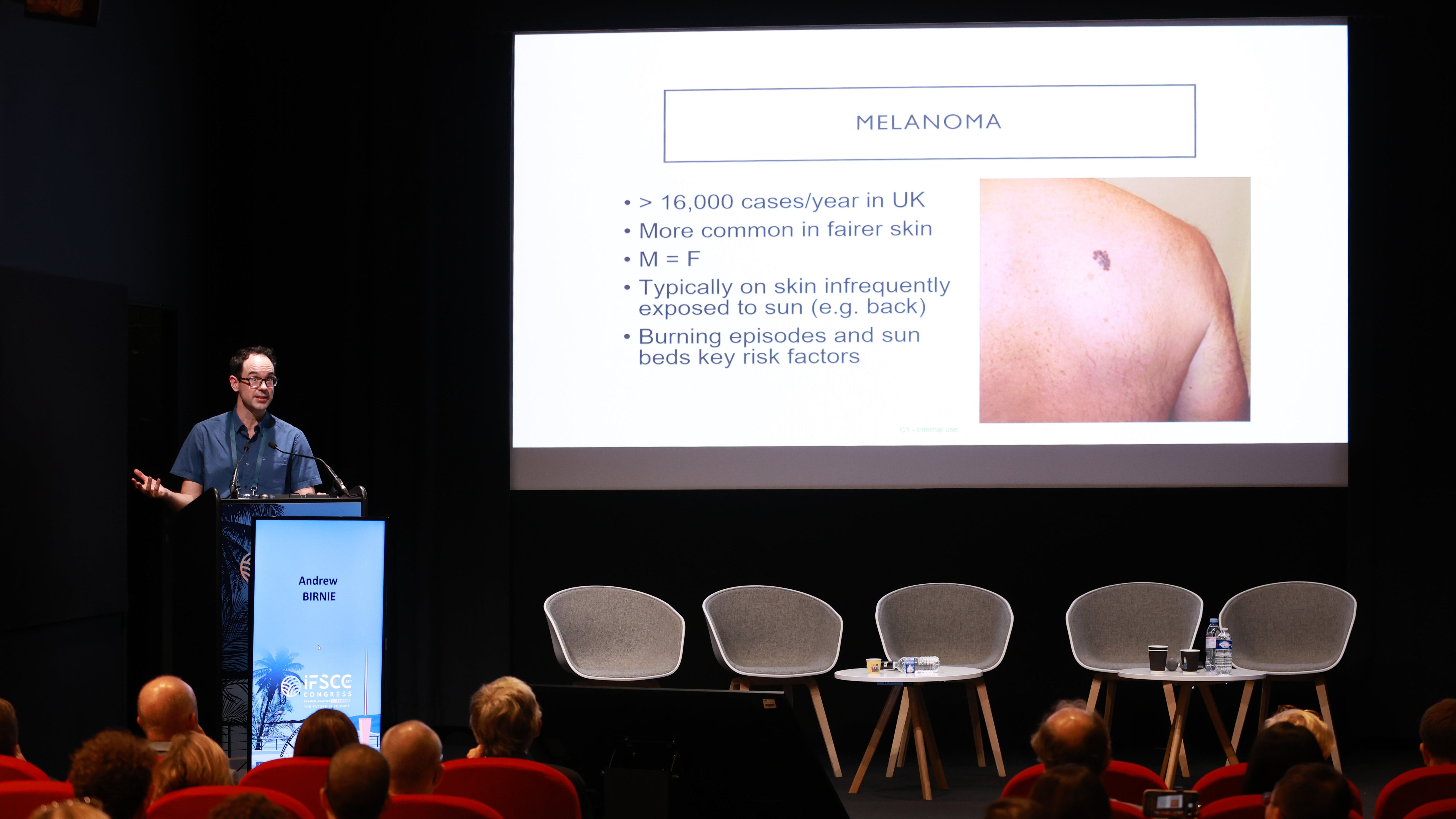 Dr Andrew Birnie at a podium with a screen displaying information about melanoma in a lecture hall.
