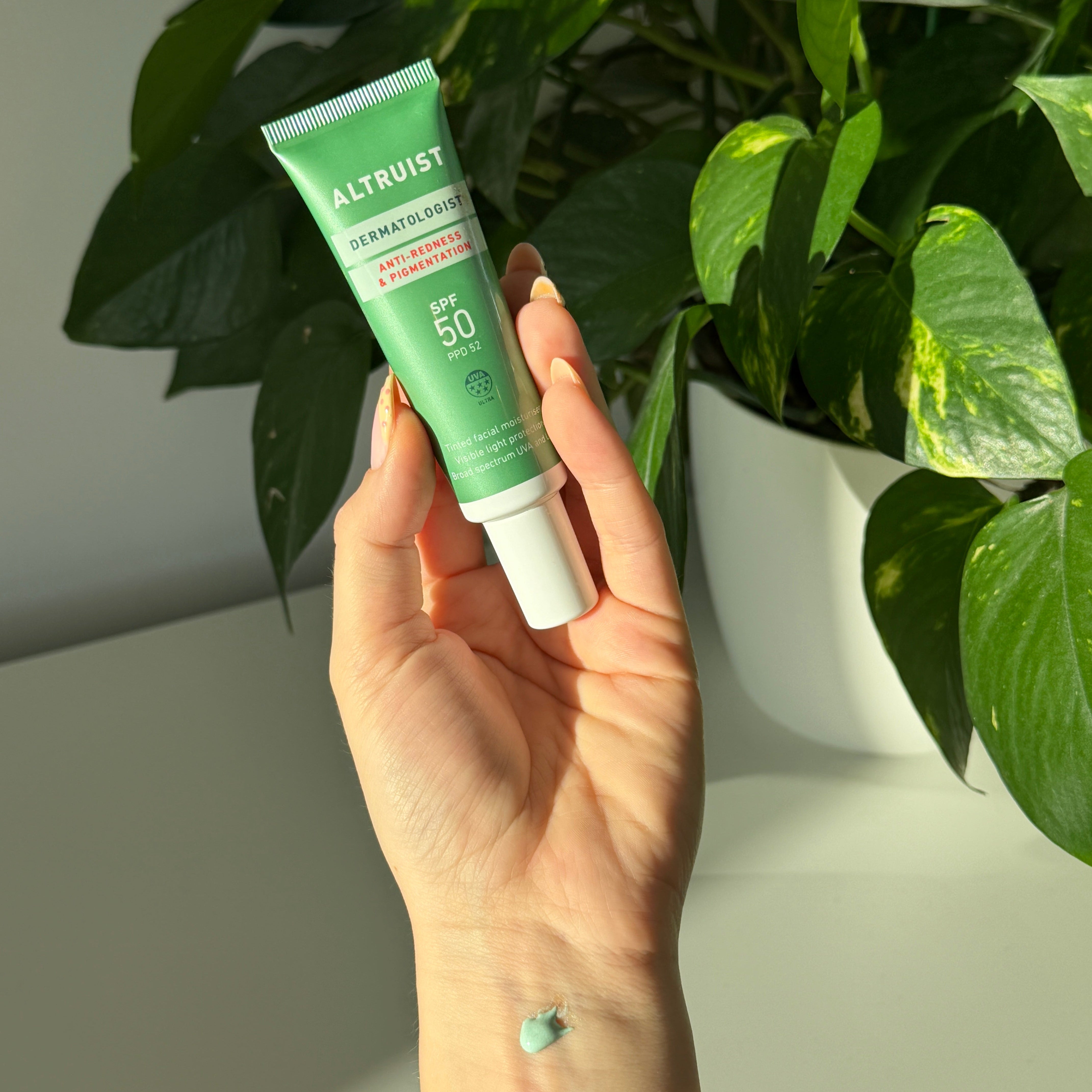 A hand holding a green tube of Altruist Anti-Redness SPF 50 against a background of green houseplants and a white wall.
