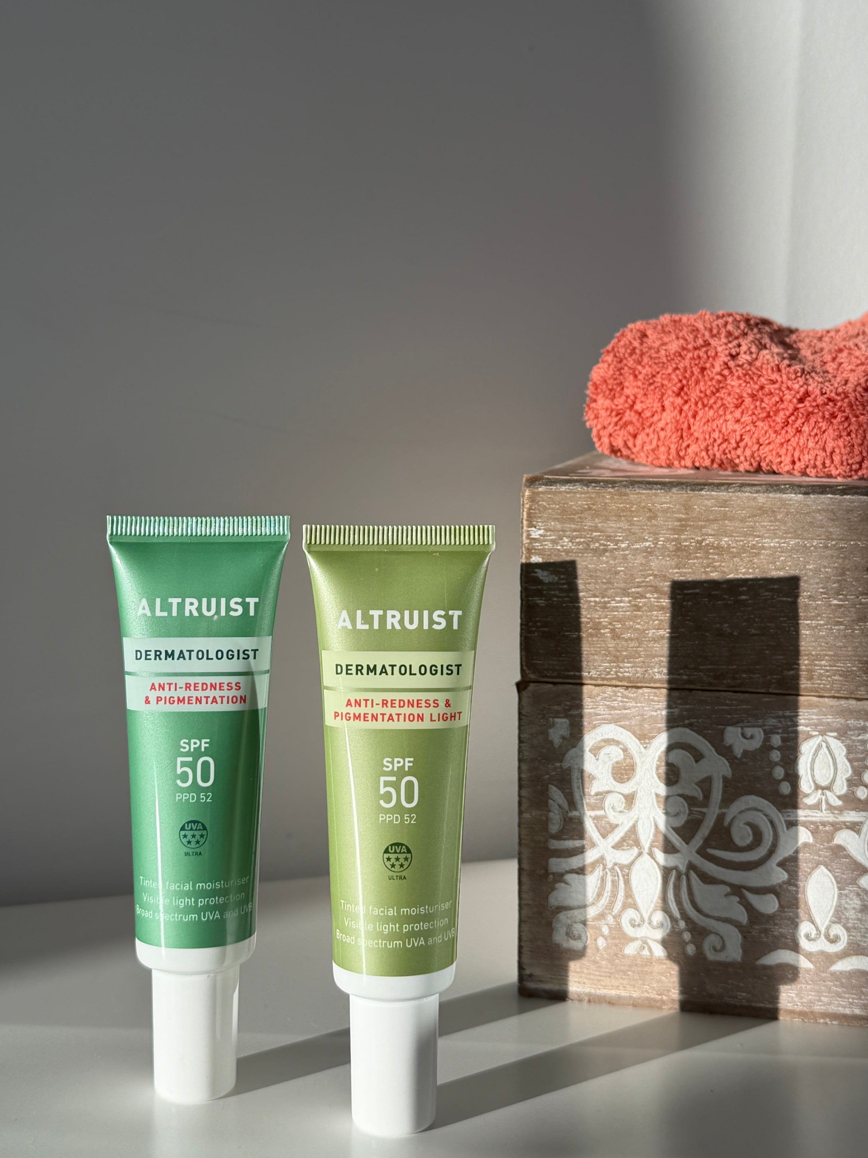 Two Altruist Anti-Redness tubes, one green and one light green, standing on a white surface next to a wooden box with a coral towel, in a styled studio setting with dramatic shadows.