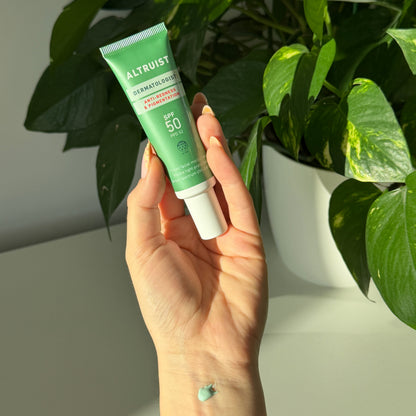 A hand holding a green tube of Altruist Anti-Redness SPF 50 against a background of green houseplants and a white wall.